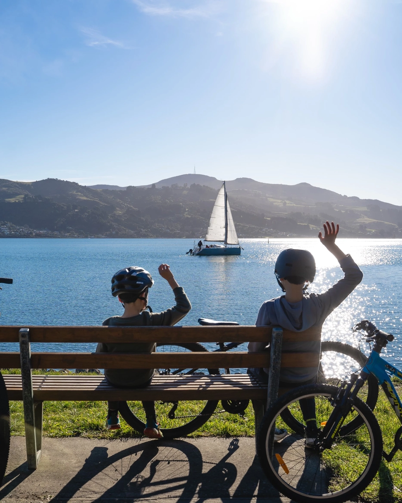 Two children with bikes at a park bench overlooking the harbour, waving at a boat. Image credit: Dunedin NZ
