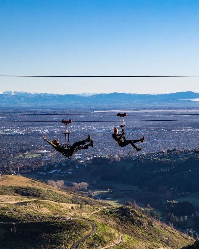 Two people ziplining at Christchurch Adventure Park with Christchurch and mountains in background, Christchurch, New Zealand.