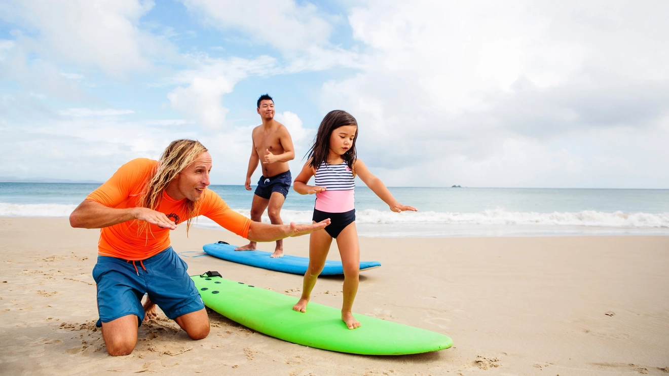 A small girl stands on a green surfboard on the sand at Clarkes Beach as an instructor kneels next to her, teaching her to surf. Image credit: Destination NSW