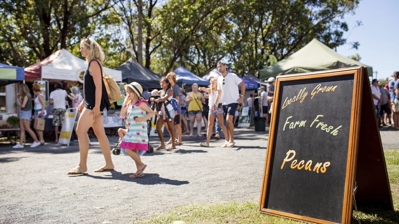 People wander the stalls of Byron Market, with a sandwich board advertising pecans in the foreground. Image credit: Destination NSW