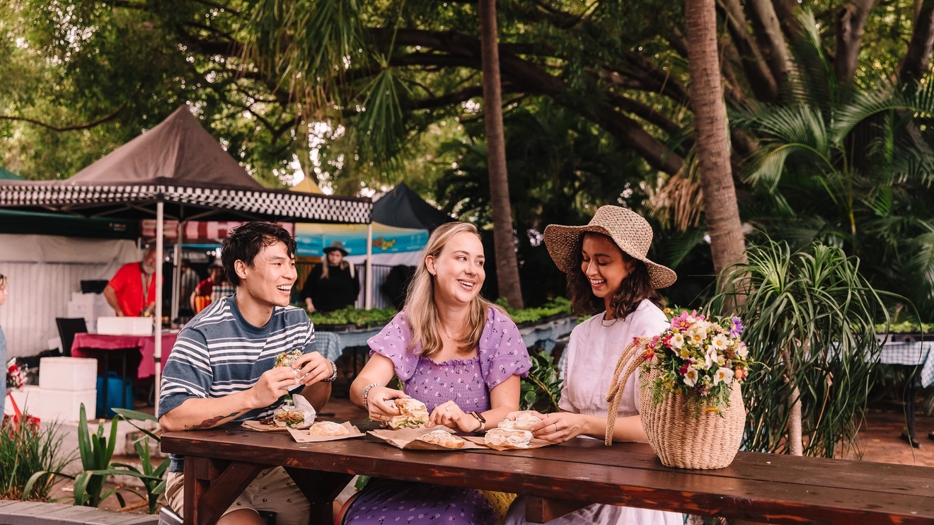 Three smiling people with sandwiches sitting at a table at Brisbane's West End Markets. Image credit: Tourism and Events Queensland