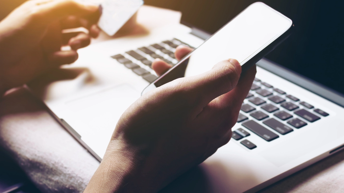 一双男人的手——一只拿着信用卡，一只拿着手机，在电脑键盘前。