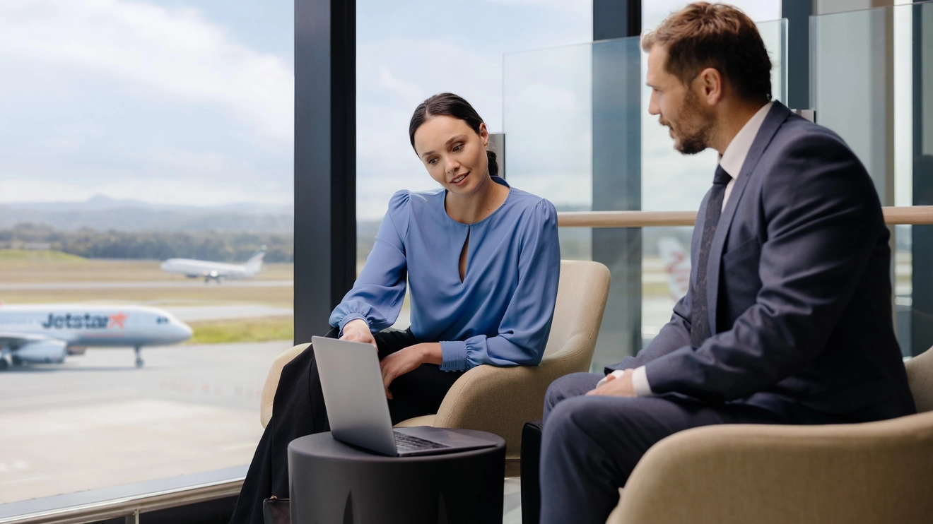 Corporate couple look at laptop in Gold Coast Airport lounge, with Jetstar plane visible through window. Image credit: Gold Coast Airport