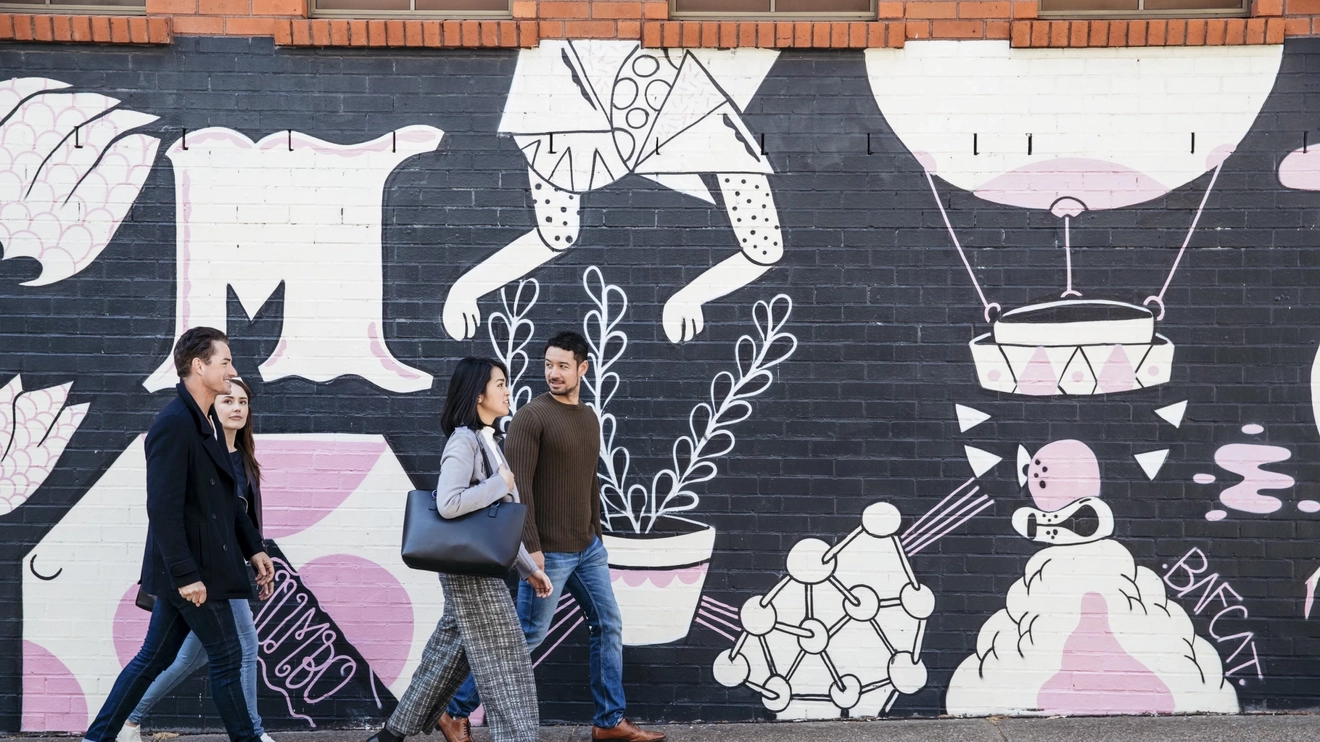 Three people walking past a wall of black, white and pink street art in Newcastle, New South Wales. Image credit: Destination NSW