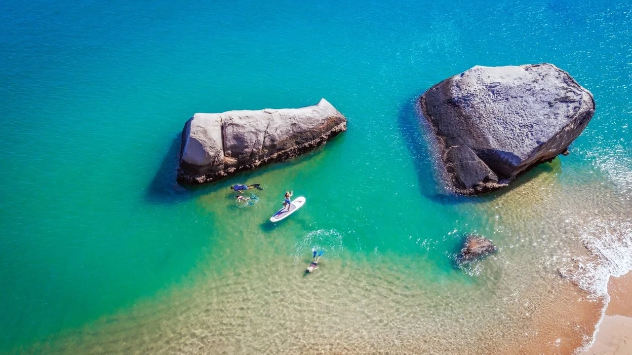 People in the water and on a SUP in clear water with large boulders in it, Magnetic Island. Image credit: Townsville Enterprise/Nathan McNeil