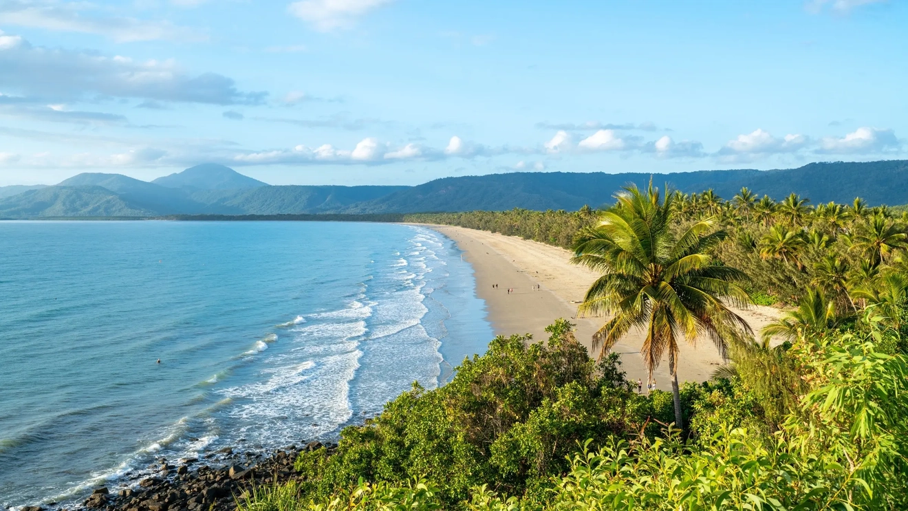 Pemandangan udara orang-orang di pantai berpasir putih dengan perairan biru dan ombak lembut, dan pegunungan di latar belakang saat matahari terbit, Port Douglas, Tropical North Queensland. Kredit foto: Tourism and Events Queensland