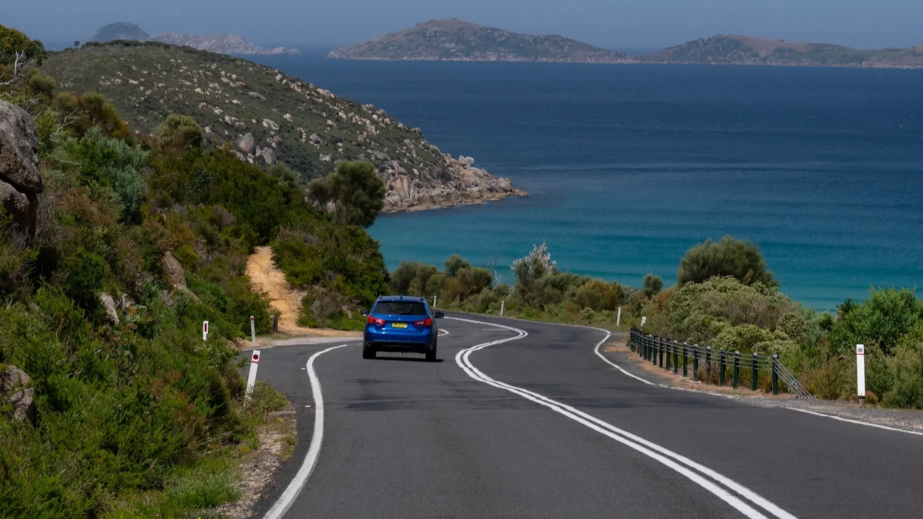 A blue car drives on the road next to a glorious blue ocean, islands on the horizon. Car rental gives you freedom to explore!