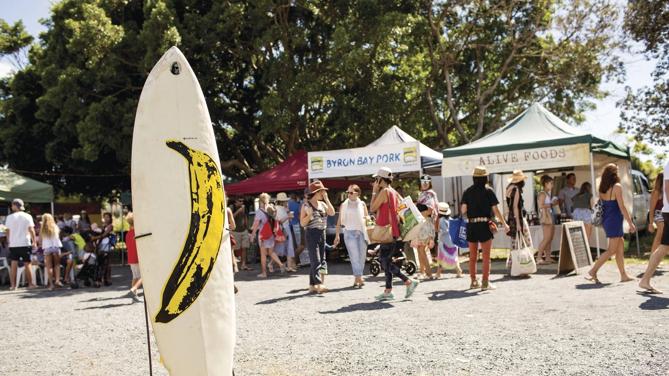 People wandering stalls at Byron Bay Market, with a surfboard sticking up out of the ground in the foreground. Image credit: Destination NSW