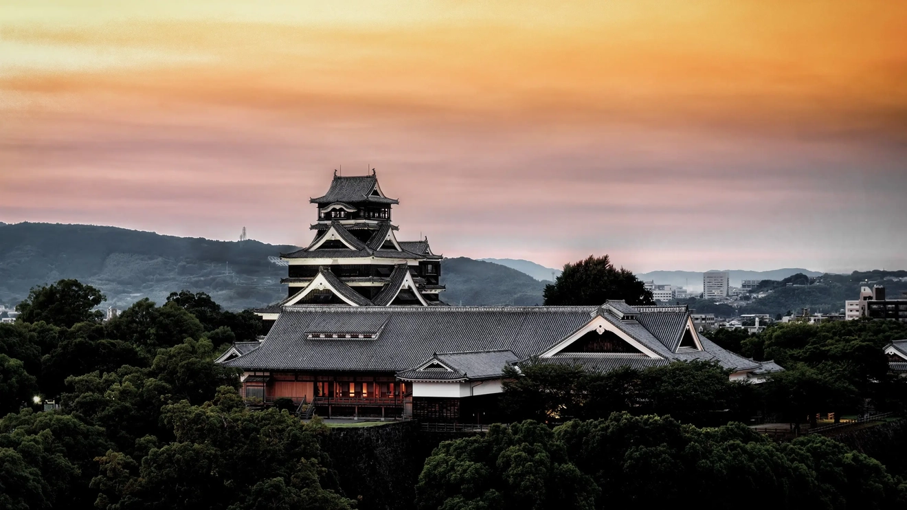 Lâu đài Kumamoto, bao quanh là cây cối, với quang cảnh thành phố Kumamoto và những ngọn núi ở phía sau lúc hoàng hôn. Kumamoto, Kyushu, Nhật Bản. Hình ảnh của: stock.adobe.com