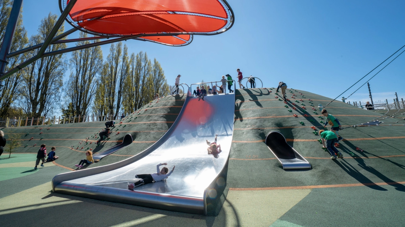 Children descending a broad slide under a red shade at Margaret Mahy Playground, Christchurch, New Zealand. Image credit: ChristchurchNZ