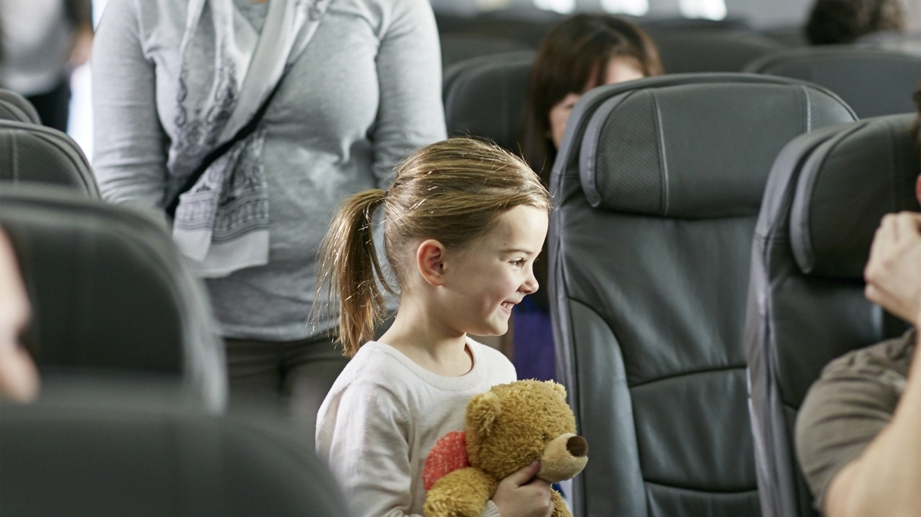 Smiling little girl stands in the aisle, about to take her seat on board a Jetstar plane. She is holding a teddy bear.