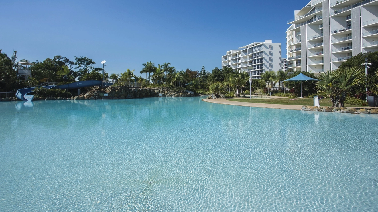 A clear blue swimming pool at Bluewater Lagoon surrounded by lawn, palms and high-rise buildings, Mackay. Image credit: Tourism and Events Queensland/Brooke Miles