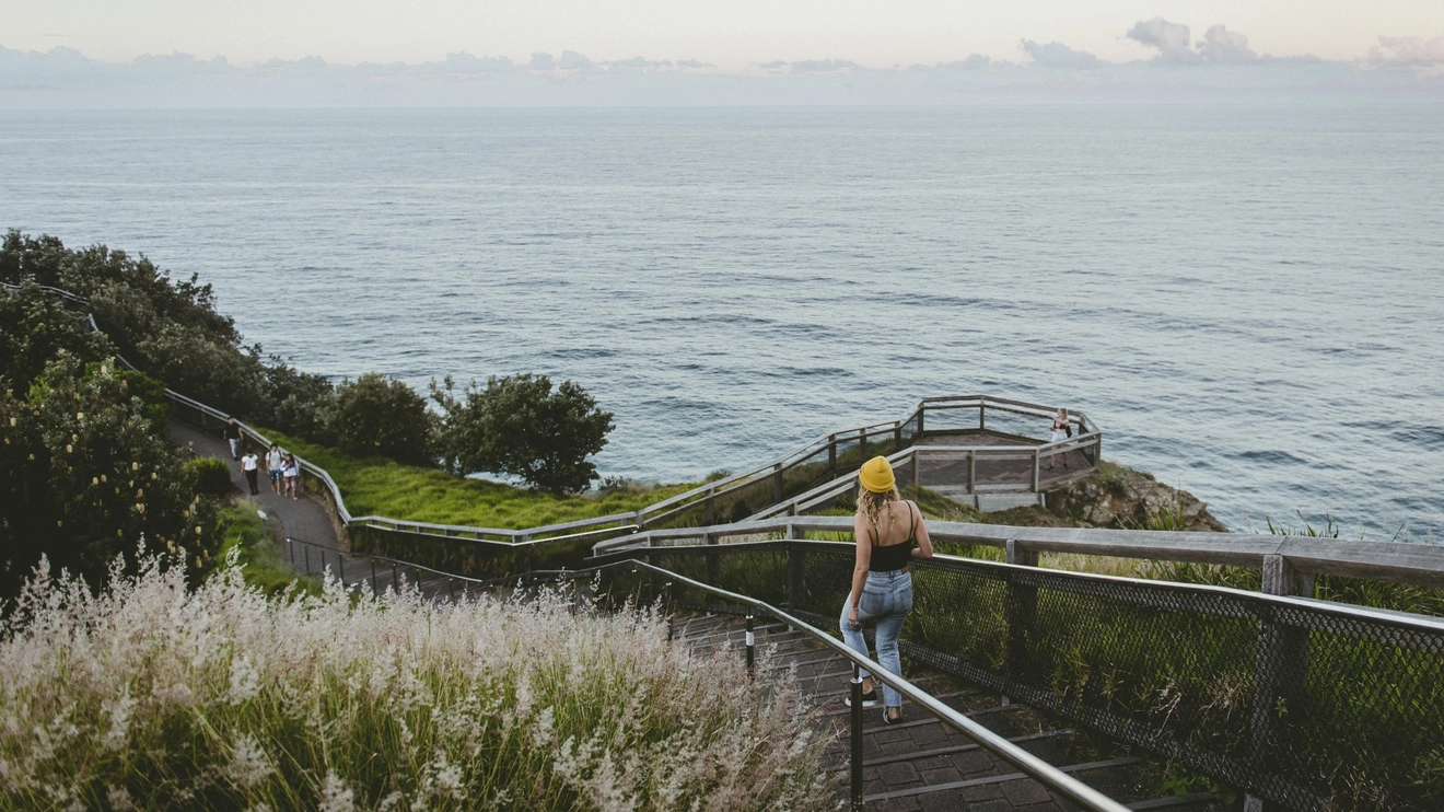 A girl walking down the Cape Byron track with plant life on the right and coast on the left. Image credit: Destination NSW